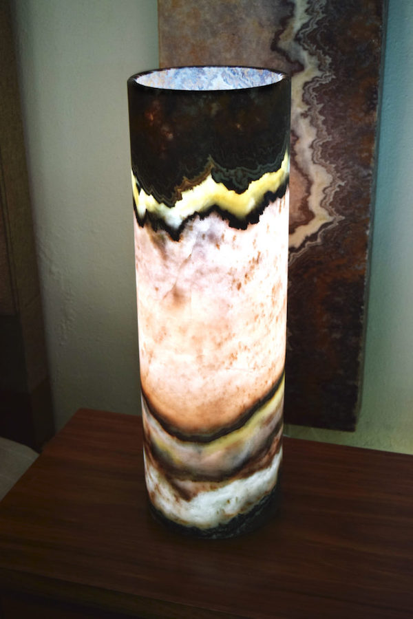 Midsize cylindrical onyx table lamp made from Beta Rosa onyx stone collection standing on the table. Top of the lamp is black, while middle part is pink, bottom - various colors.