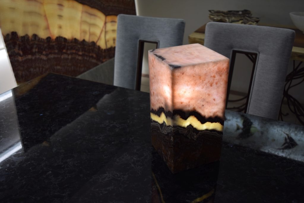 Small-sized rectangular table lamp standing on the dinner table and made from Beta Rosa onyx stone collection. The top of the lamp is pink while the bottom is black and horizontal yellow line in the middle. The light is on.