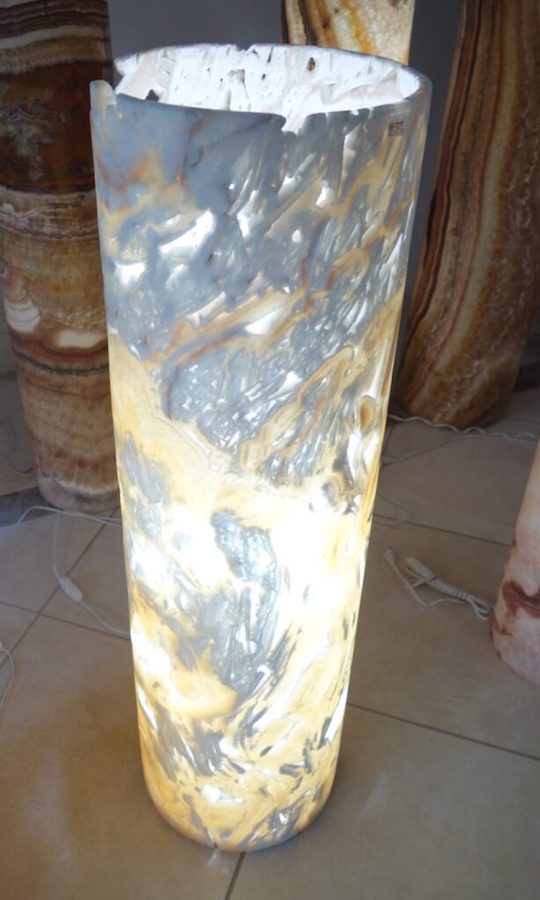 Onyx Marble Cylindrical Floor Lamp with Open Curved Top and Light On. Picado Collection. Colors: Various Shades of Yellow and Gray. Picado Onyx Marble has Natural Holes.