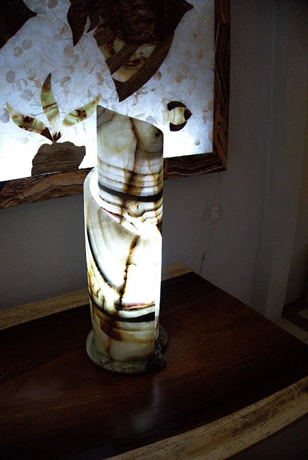 Spiral onyx table lamp made from Onyx Verde Talan stone collection standing on the nightstand with light on. Main colors are white, brown, black, and green